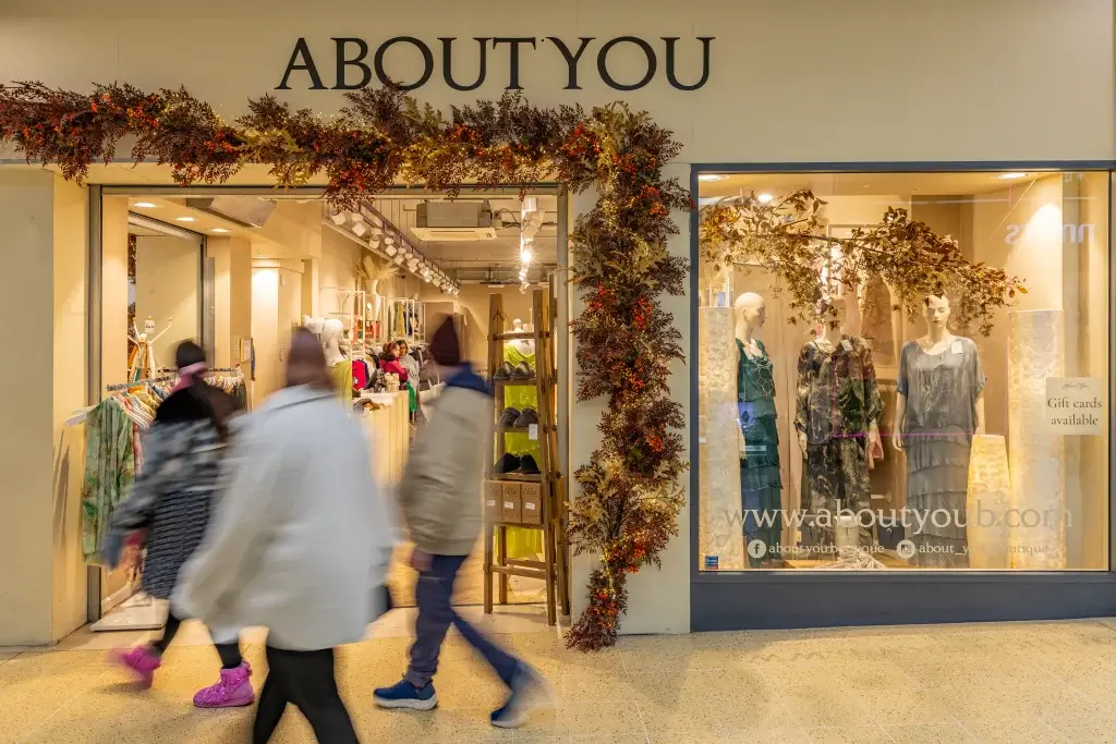 A busy storefront of "ABOUT YOU" adorned with autumn decor, featuring mannequins in stylish outfits and shoppers passing by.