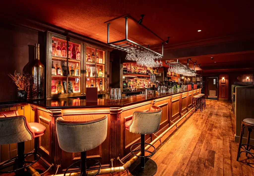 Warm, dimly lit pub interior with a long polished wooden bar, upholstered bar stools and backlit bottle shelves.