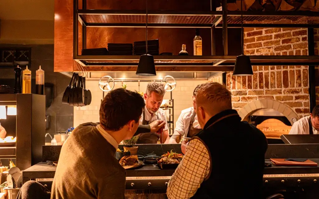 Two diners at a restaurant counter watch chefs plating dishes in a warmly lit open kitchen with a brick oven.