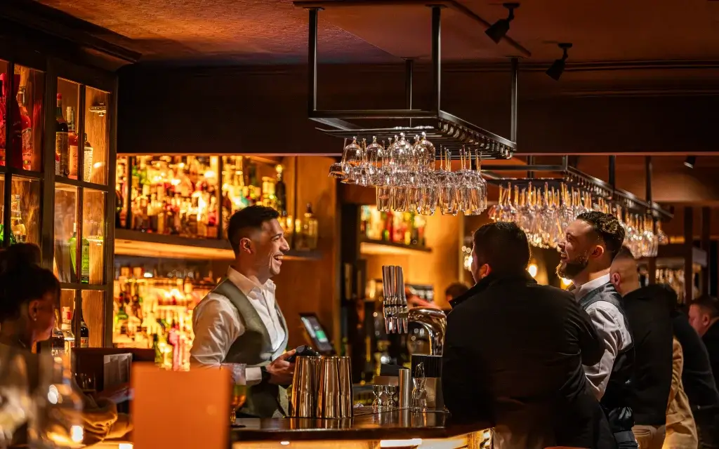 Bartender laughing with two male patrons at a warmly lit bar, rows of hanging wineglasses and backlit bottles behind them.