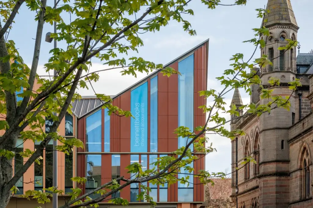 Red-brown and blue-glass office with vertical bennettbrooks signage, framed by green tree branches and a stone church tower.
