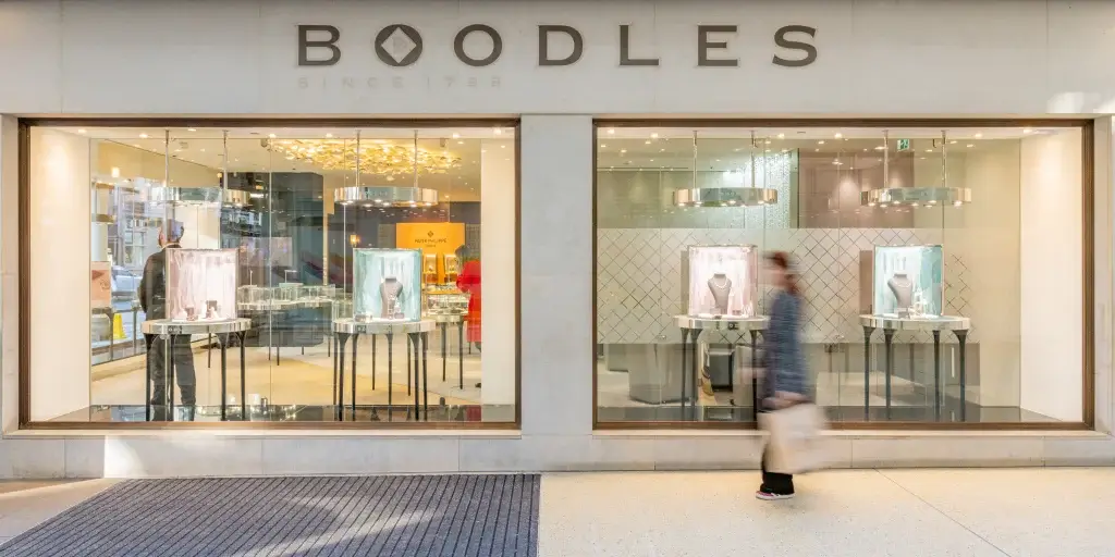 A blurred figure strolls past a Boodles jewelry store window displaying elegant pieces and modern decor.