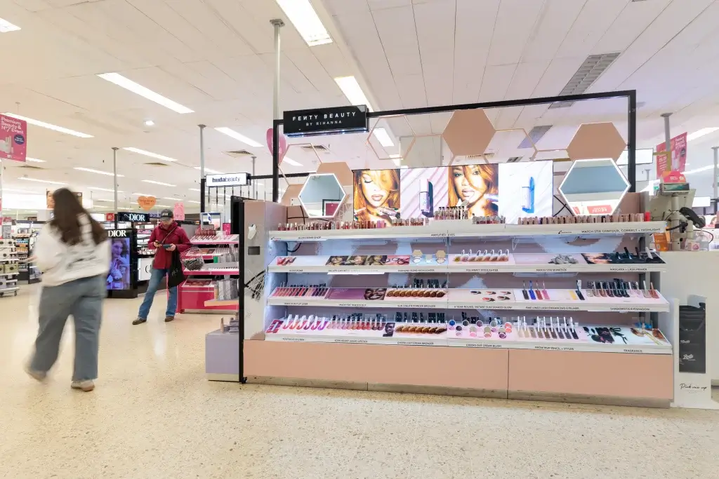 Fenty Beauty counter in a department store with illuminated screens of model faces, organised shelves of lipsticks and makeup and shoppers nearby.