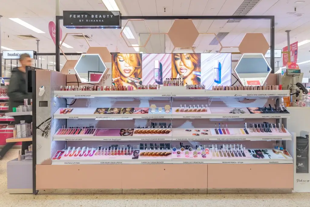 Fenty Beauty cosmetics display with hexagonal mirrors, shelves of lipsticks, palettes and a large screen showing a model.