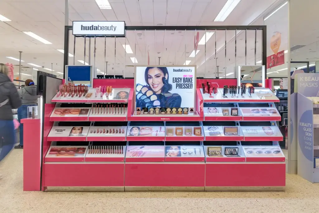 Huda Beauty pink cosmetics display in a store with shelves of makeup and central "Easy Bake Pressed" ad with smiling model