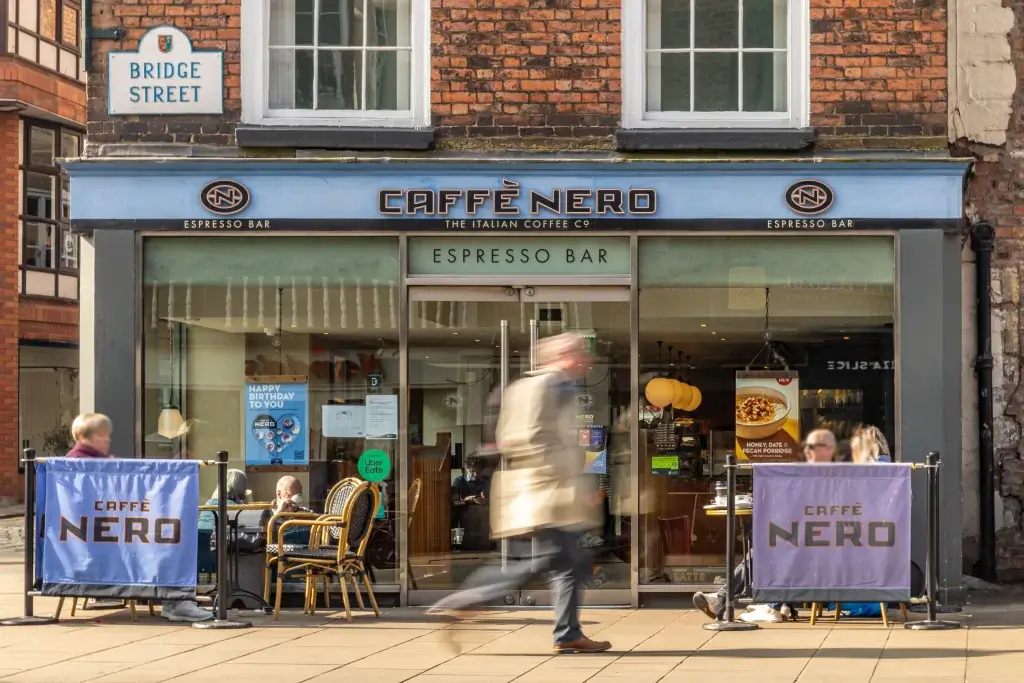 A bustling Caffè Nero coffee shop entrance with outdoor seating, inviting signs, and patrons enjoying their time. Bright sunlight illuminates the scene.