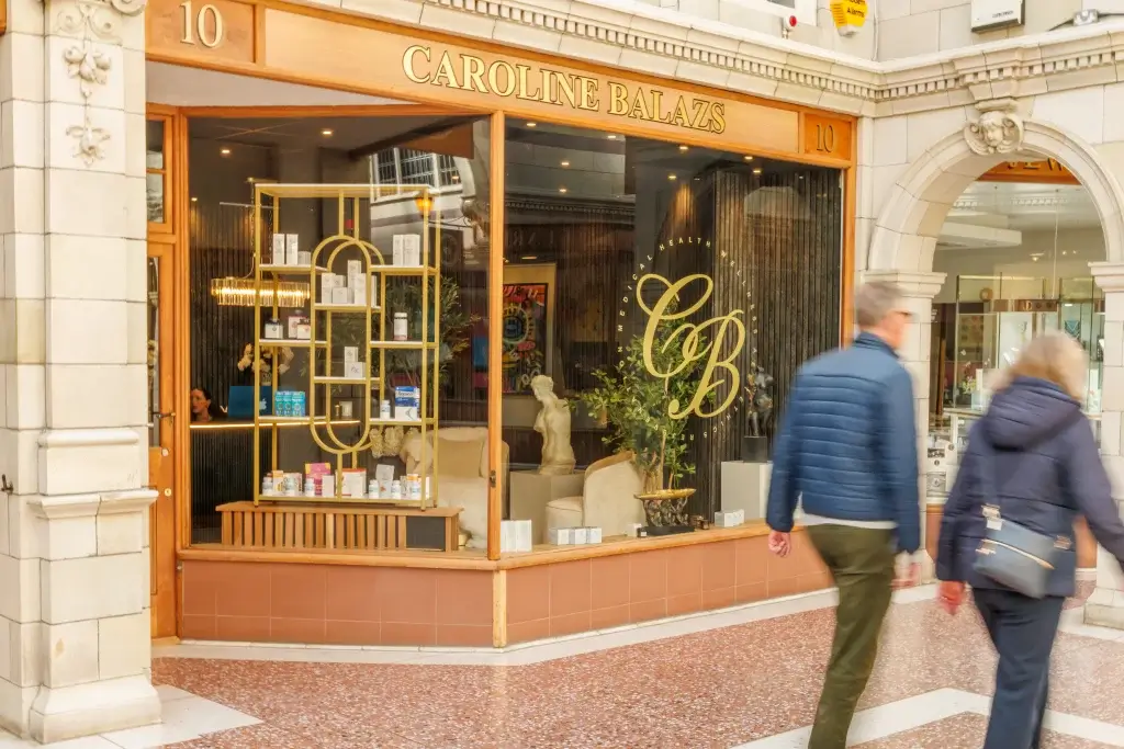 Caroline Balazs boutique storefront with gold "CB" logo and product-filled display shelf visible through the window, two blurred shoppers passing by