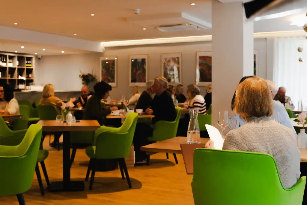 A bustling restaurant scene with patrons seated at wooden tables, surrounded by vibrant green chairs and artistic wall decorations.