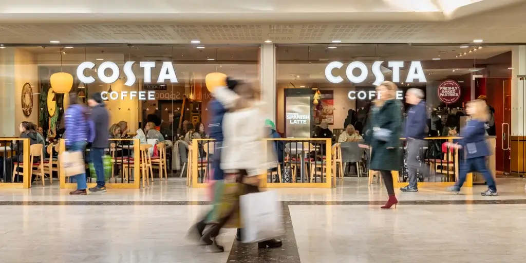 A bustling Costa Coffee shop with patrons inside while shoppers walk by, showcasing a lively café ambiance in a shopping area.