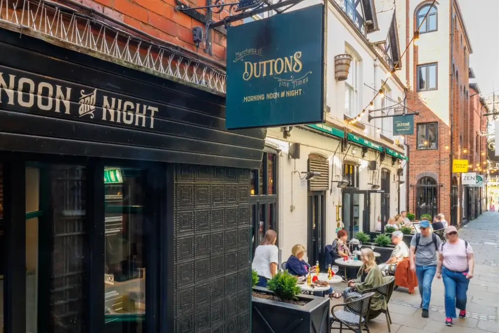 Outdoor street café on a narrow brick lane with diners under string lights and a hanging sign reading Duttons