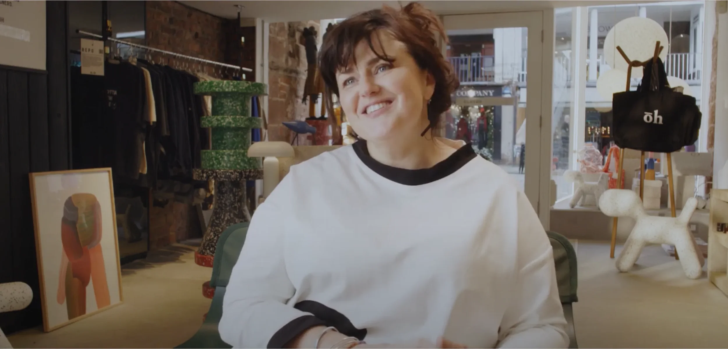 Uná Meehan, Co Lead of Good For Nothing Chester in a white shirt sitting in a shop with artwork and clothes on display.