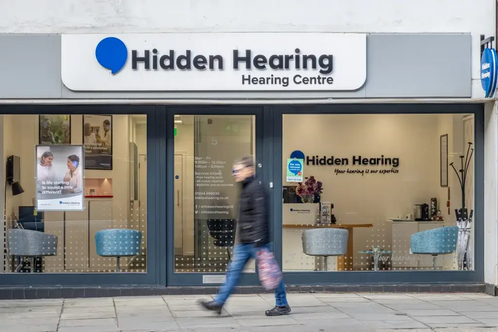 A person walks past the Hidden Hearing Centre, featuring modern exterior design with seating and informative posters displayed.
