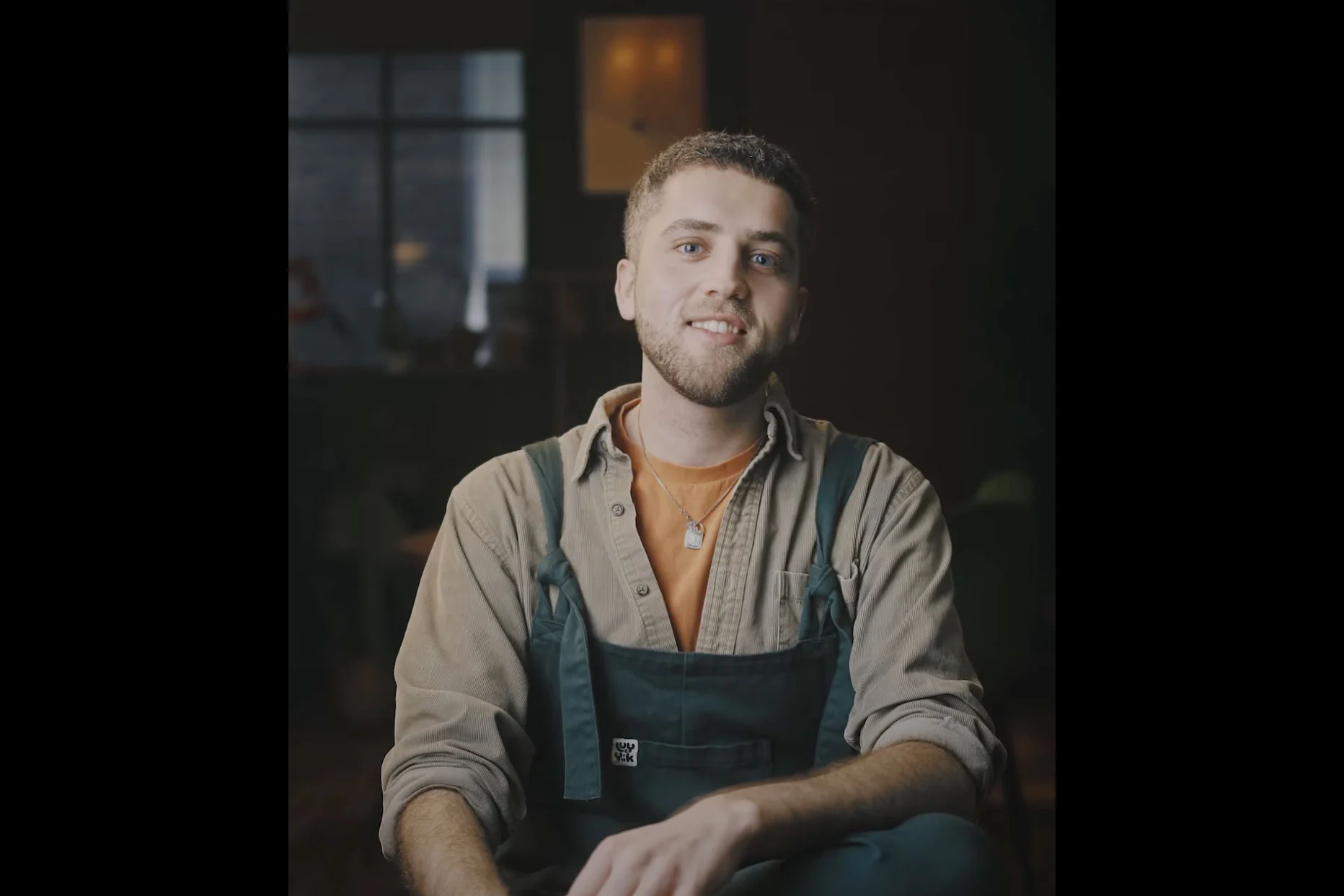 Ian from Chester BID wearing overalls and a casual shirt, smiling confidently while seated in a warmly lit room.