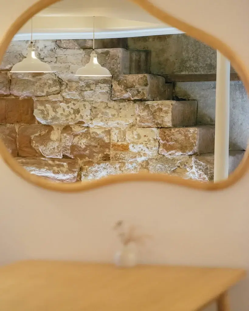 A curved mirror reflects rustic stone stairs and two pendant lights, creating a warm, inviting atmosphere. A small vase with dried flowers sits below.