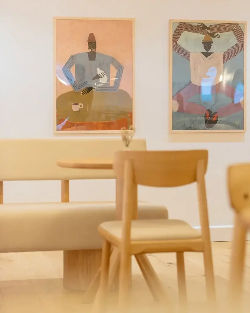 A cozy café interior featuring light wooden furniture and two colorful framed artworks on the wall, enhancing the inviting atmosphere.