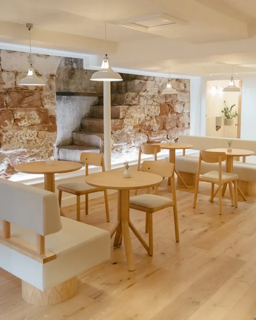 A modern cafe interior featuring wooden tables, chairs, and light fixtures, with a rustic stone wall and soft seating.