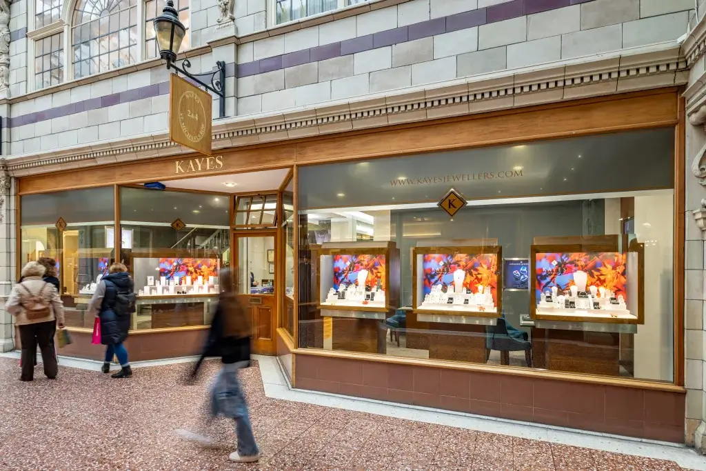 A jewelry store window display featuring elegant jewelry pieces, with shoppers walking by in a vibrant shopping arcade.