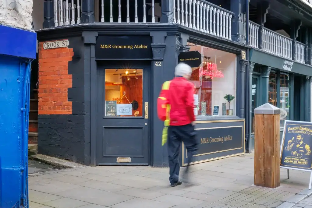 Black corner shop M&R Grooming Atelier with neon window display and a blurred man in a red jacket walking past.