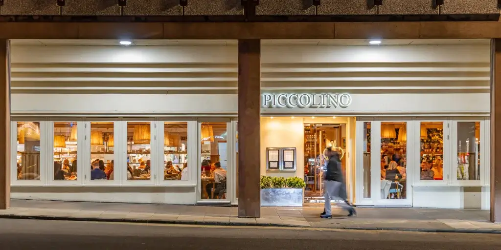 Exterior of Piccolino restaurant at night, showcasing warm indoor lighting, diners visible through large windows, and a passerby.