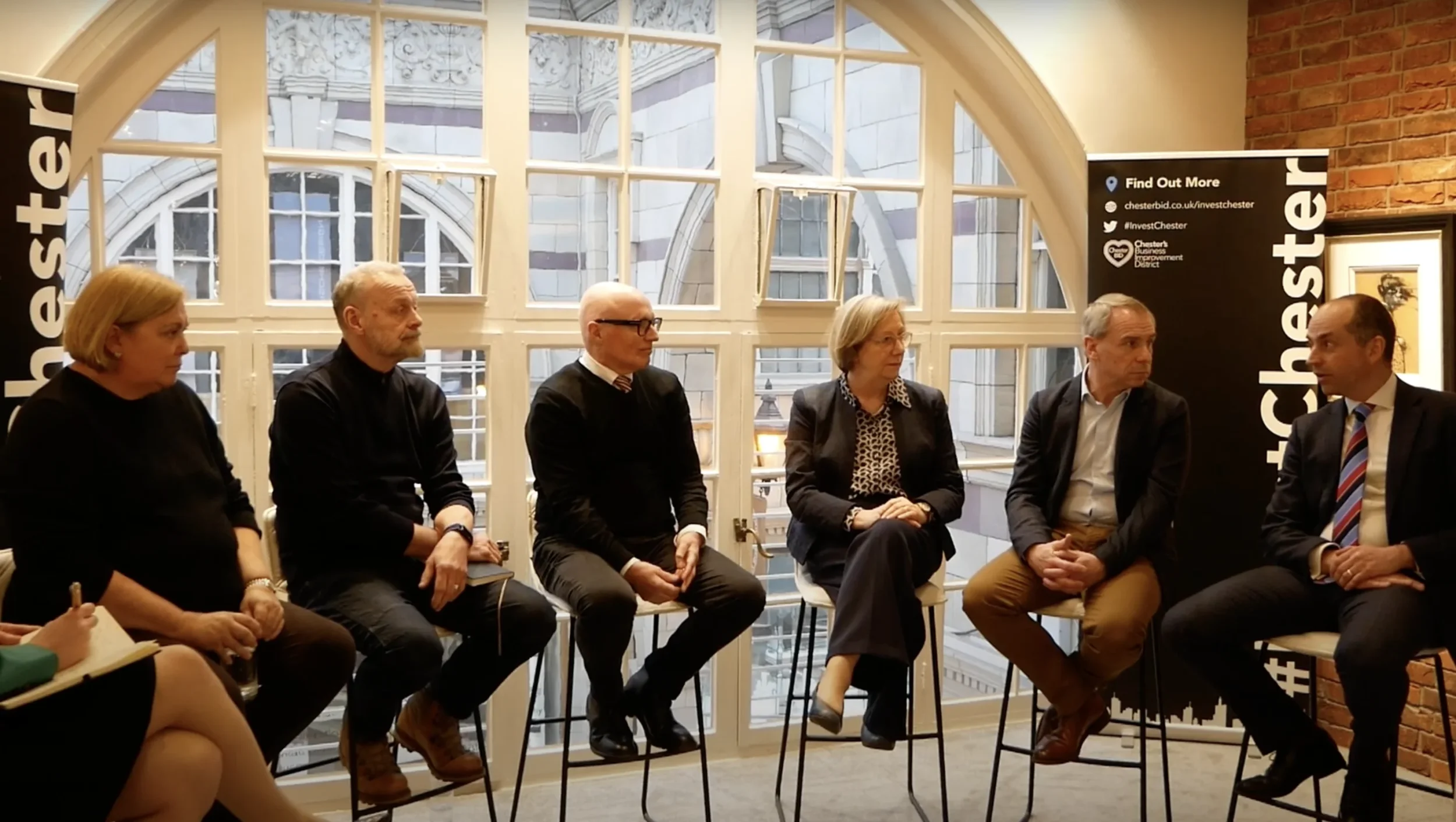 A group of individuals seated for a Place North West panel discussion in a room with large windows.