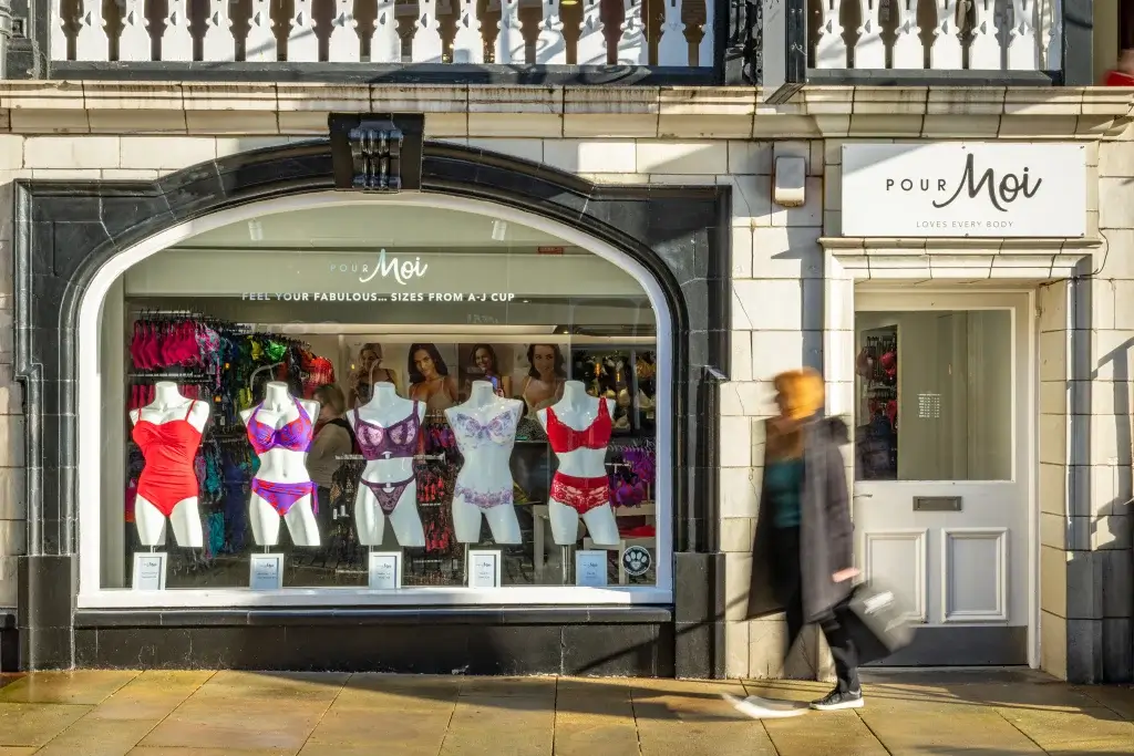 A storefront displaying colorful lingerie on mannequins, with the sign "Pour Moi" above and a blurred passerby in front.