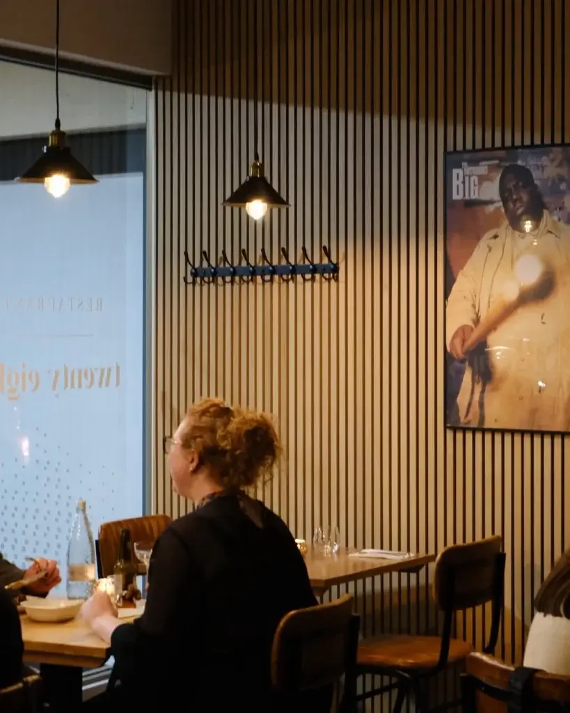 A cozy restaurant interior with wooden slats, pendant lights, and a poster on the wall while diners enjoy their meals at wooden tables.
