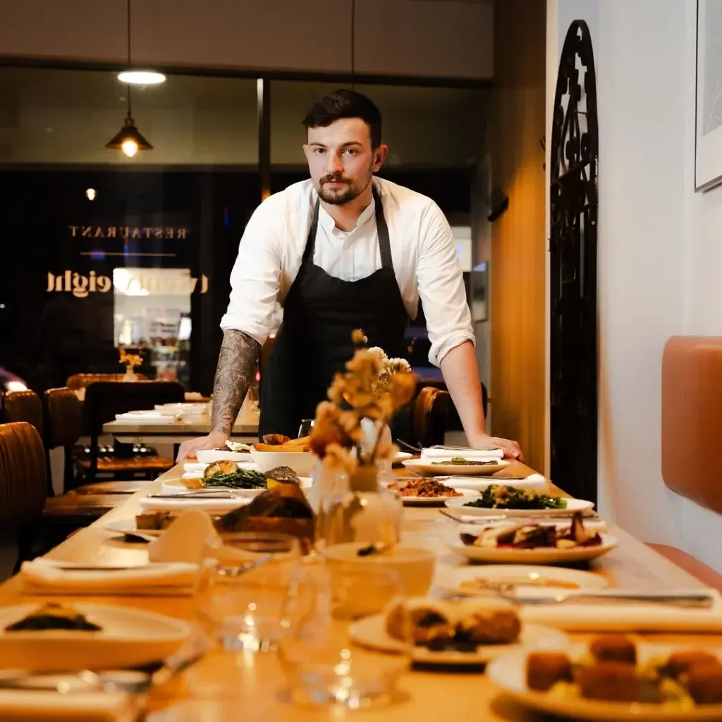 A chef stands at a beautifully set dining table filled with various gourmet dishes and elegant tableware, exuding a warm dining atmosphere.