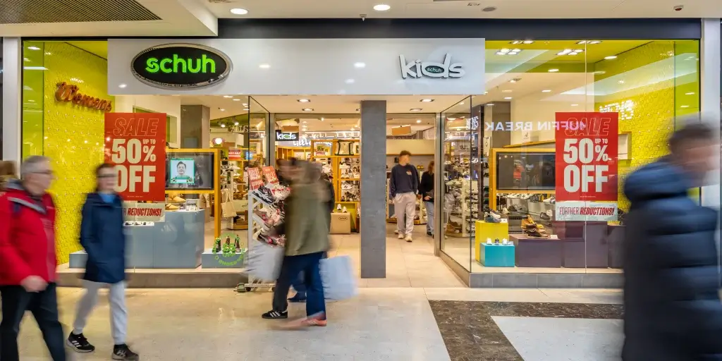 Busy shopping mall entrance of a schuh store featuring a "50% OFF" sale sign, with shoppers passing by.