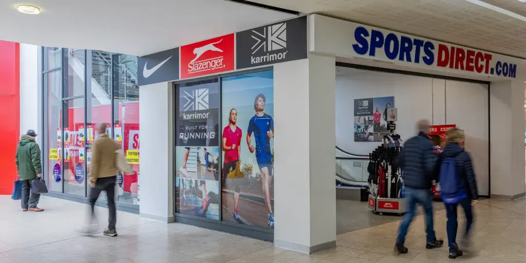 Entrance of a Sports Direct store featuring advertisements for Karrimor and Slazenger, with shoppers walking by.
