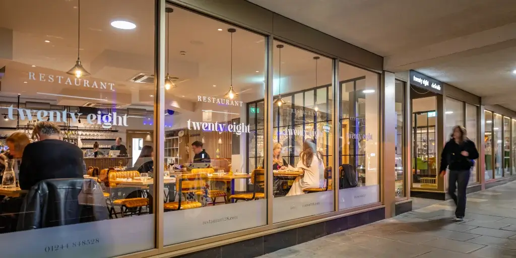 Illuminated restaurant "twenty eight" with diners visible through large windows, as a passerby walks by on a city street at night.