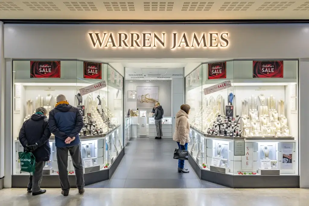 Exterior view of a Warren James jewelry store, featuring illuminated displays and customers browsing during a sale event.