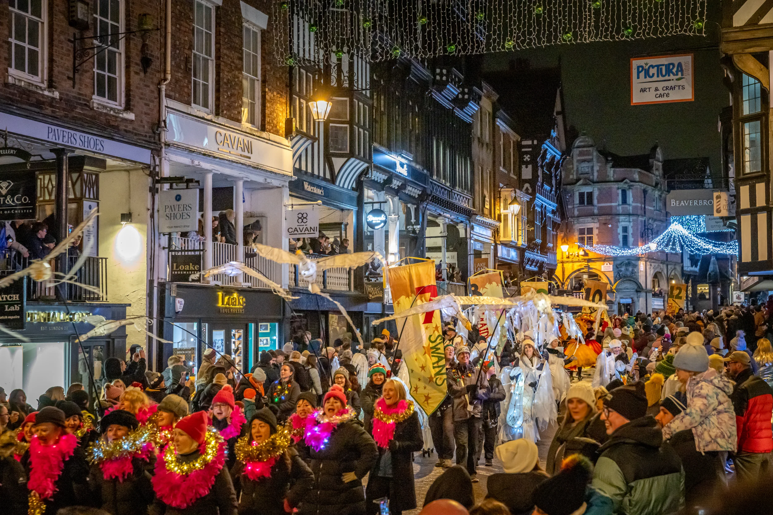 Night-time street parade with performers in pink tinsel collars, glowing white swan puppets and banners amid festive lights.