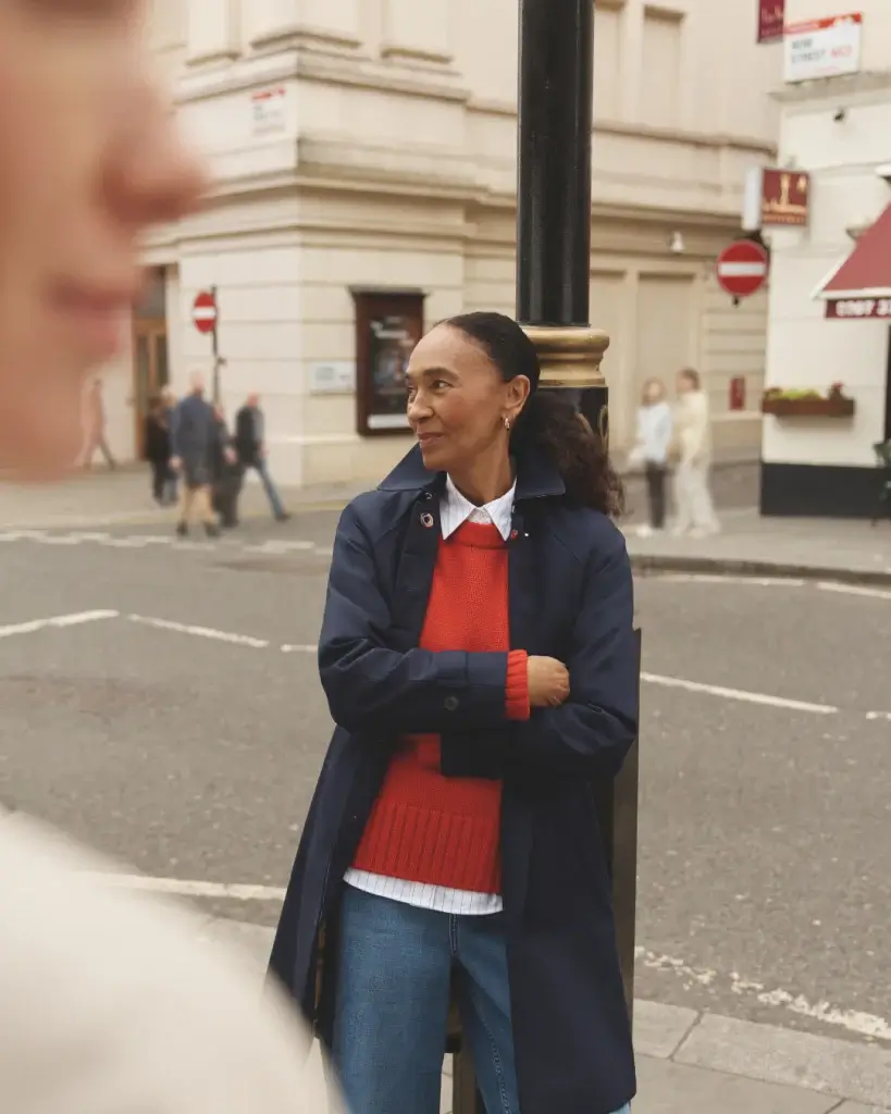 Woman leaning against a lamppost at a street corner wearing a navy coat, red sweater, white shirt and jeans.