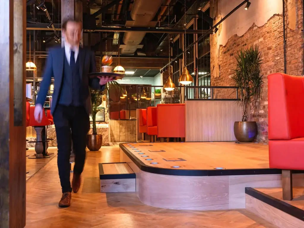 A blurred waiter in formal attire carries a drink on a tray through a modern restaurant with rustic decor and vibrant red seating.