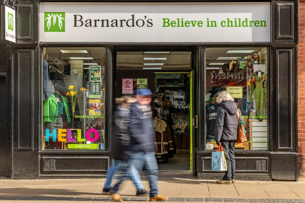 A busy storefront of Barnardo's charity shop, displaying colorful signs and clothing, with shoppers passing by in Chester.