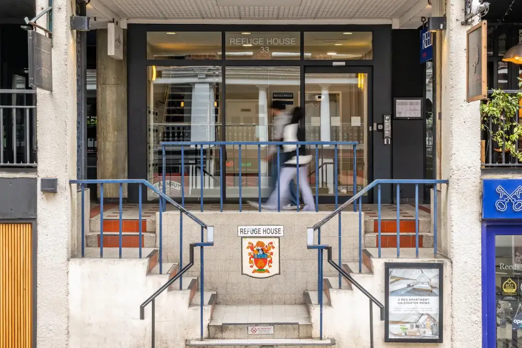 Two people walk past the entrance of Refuge House, a modern glass-fronted building with a blue railing and visible elevator.