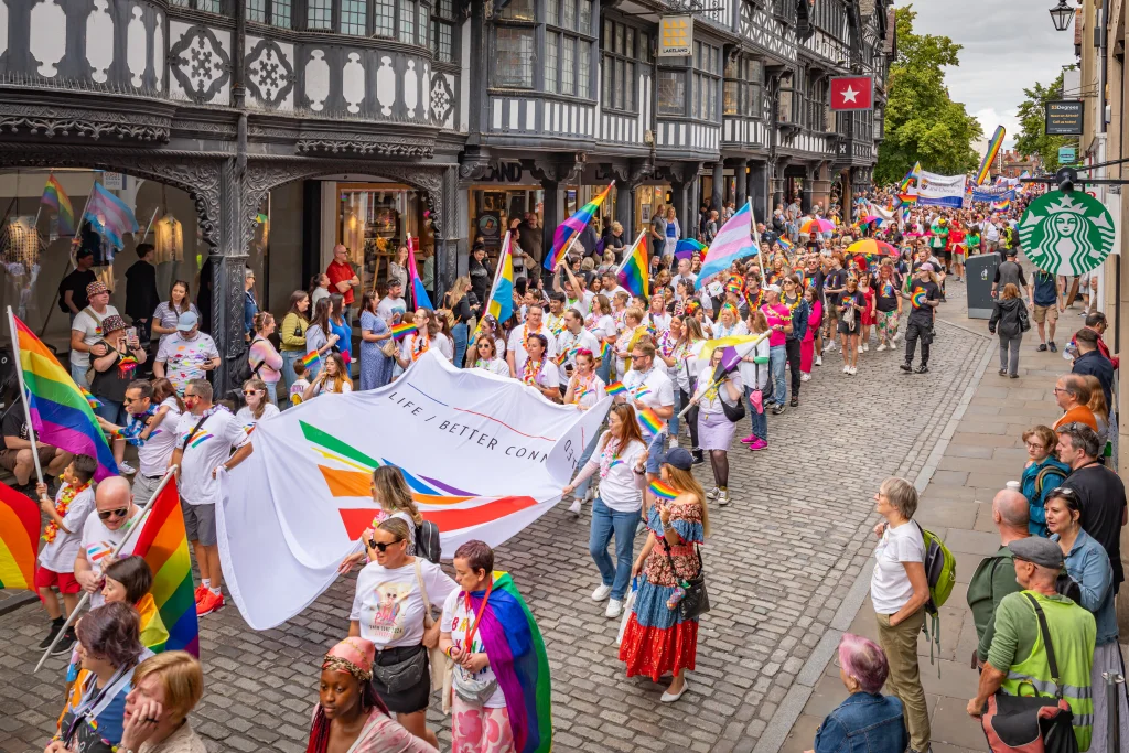 Crowded Pride parade on a cobbled street with marchers waving rainbow and trans flags and a large banner, spectators watching.