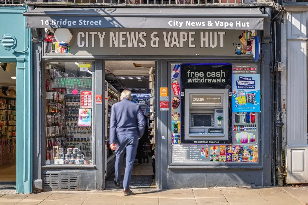 A man in a suit enters the City News & Vape Hut, featuring an ATM and vibrant displays of drinks, vape products, and snacks.