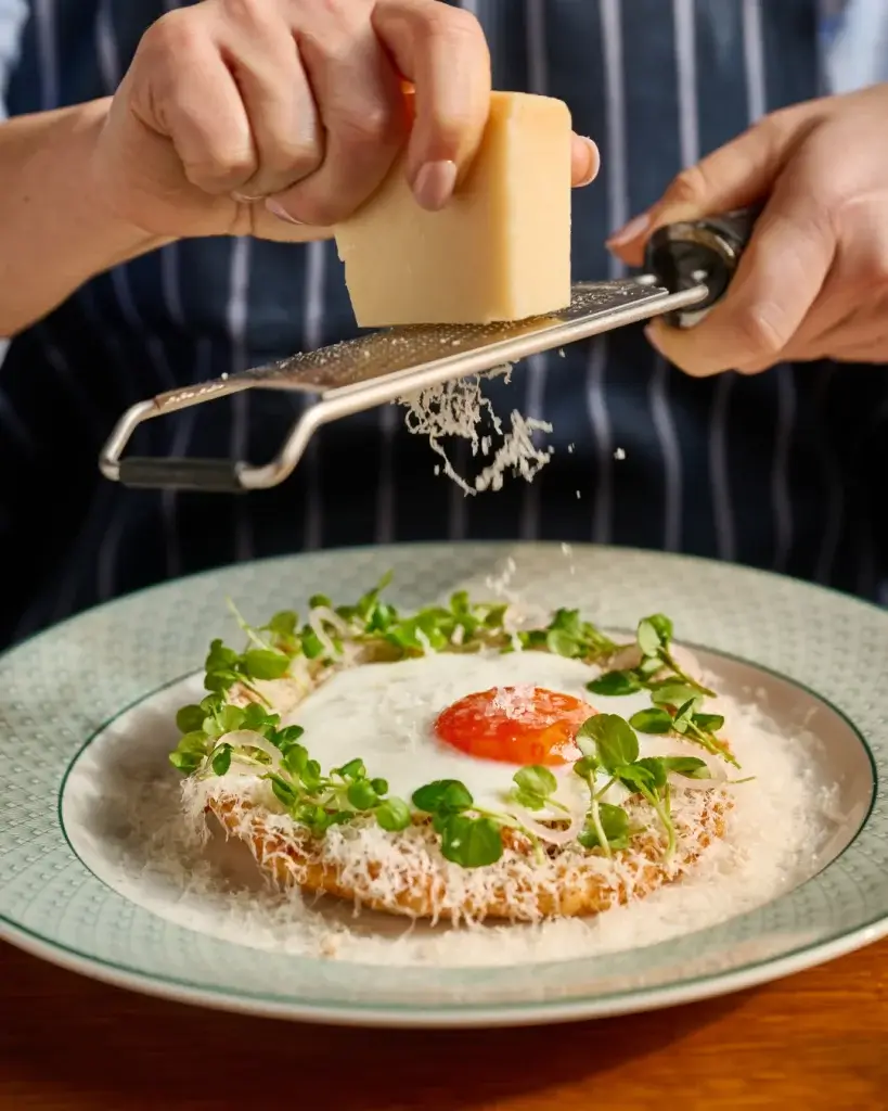 Hands grating hard cheese with a microplane over a fried-egg-topped toast ring garnished with microgreens.