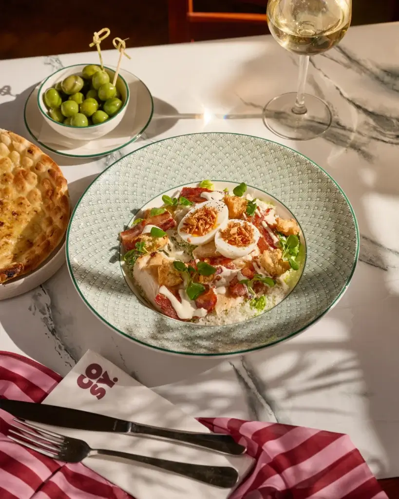 Salad with halved boiled eggs, bacon, croutons and creamy dressing in a patterned bowl, with olives and a glass of white wine.
