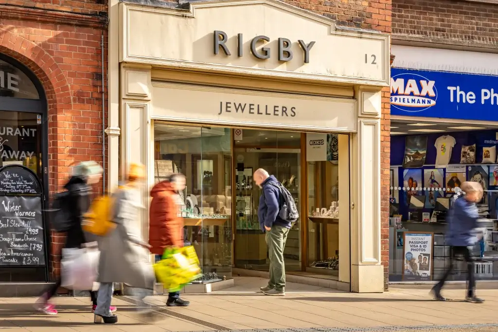A busy street scene featuring Rigby Jewellers, with shoppers passing by carrying bags, against a backdrop of varied storefronts.
