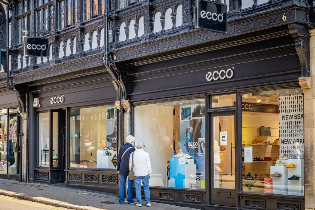Two people admire shoe displays in the storefront of an Ecco shop, highlighting products and sale promotions.