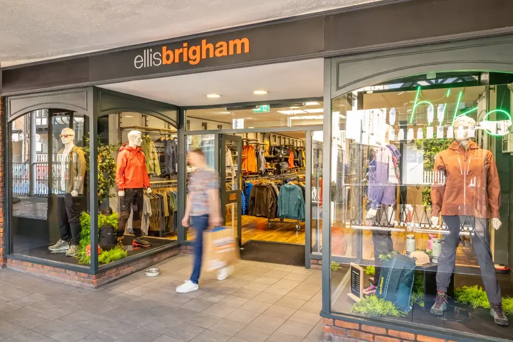 The entrance of an outdoor gear store showcasing mannequins in colorful clothing, with a customer walking out carrying a shopping bag.