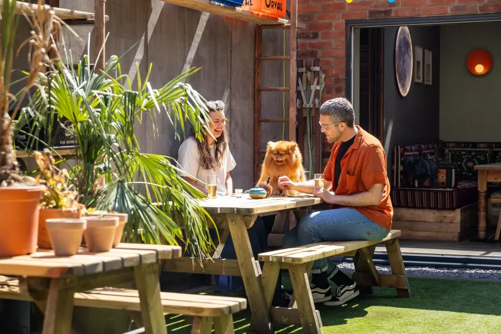 A sunny outdoor café scene featuring two people enjoying drinks with a fluffy dog on a wooden table surrounded by greenery.