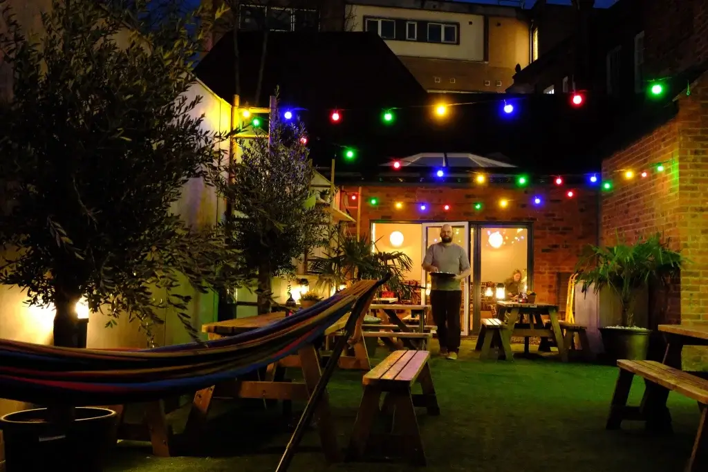 A cozy outdoor area with colorful lights, wooden picnic tables, and lush greenery, creating a warm evening atmosphere.