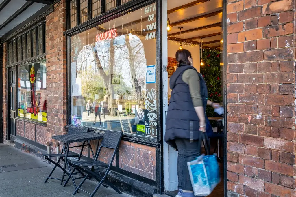 A woman wearing a dark vest enters a café named "Gaudi's," with colorful signage and a cozy interior visible through the window.