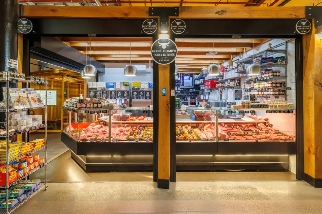 Butcher shop front with Geoff Hughes Family Butcher sign, glass display cases filled with assorted meat cuts and jars.