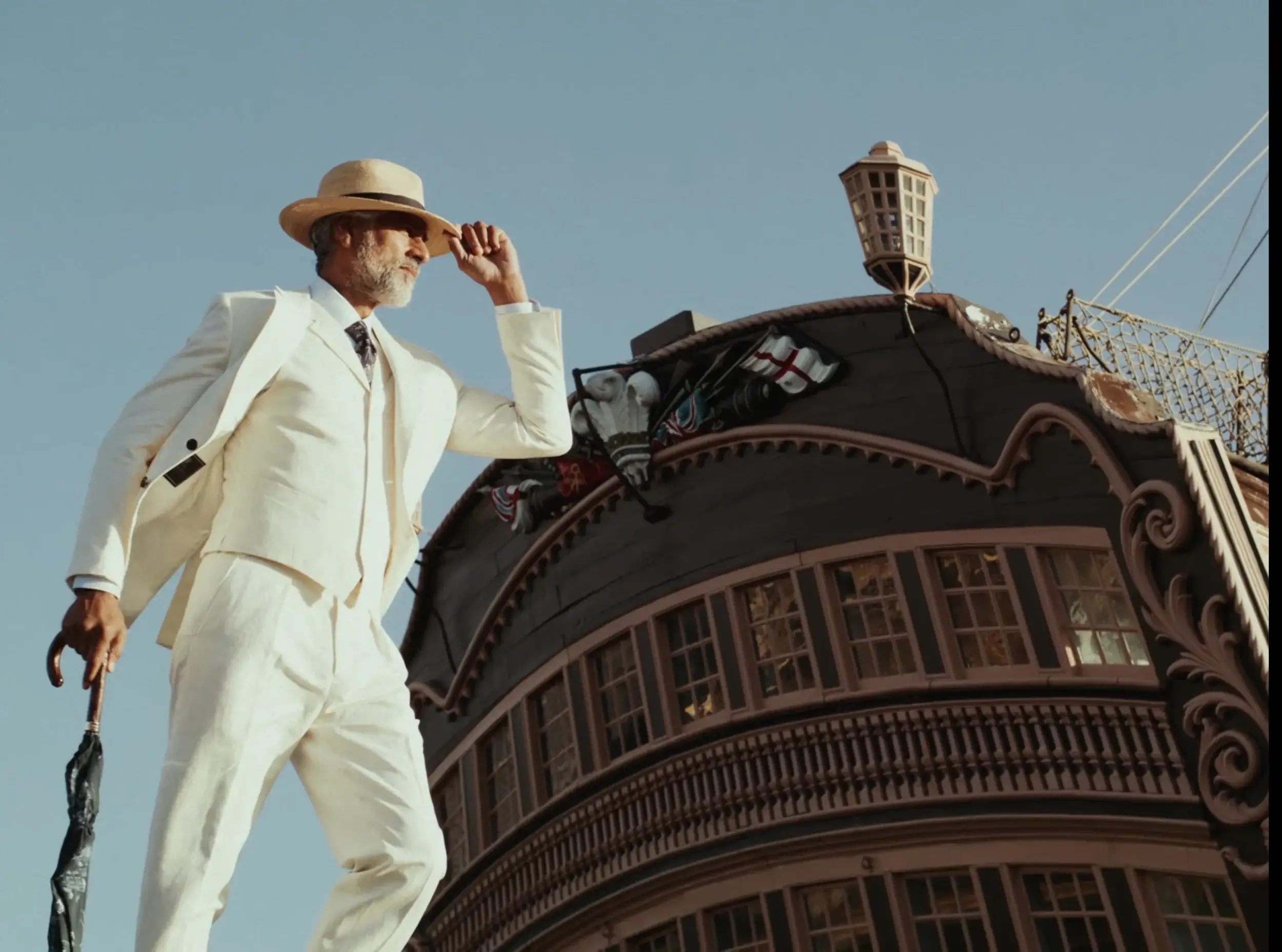 Bearded man in a white suit and straw hat tipping his hat and holding an umbrella beside an ornate ship stern.
