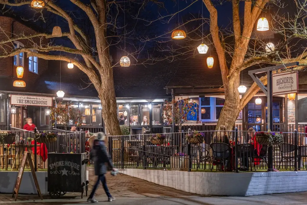 Cozy outdoor dining at The Smokehouse, adorned with lanterns, surrounded by trees, and bustling with patrons enjoying the evening ambiance.