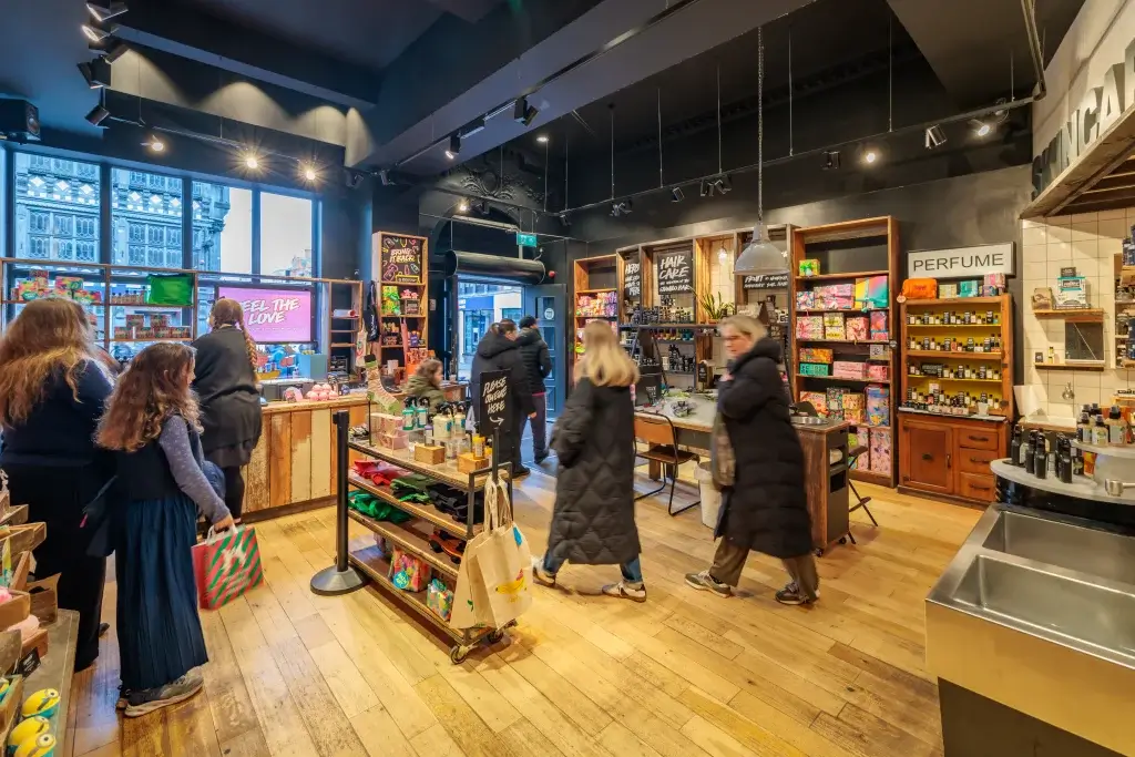 Busy boutique store interior with wooden floors, shelves of colourful cosmetics and perfume, and shoppers browsing.